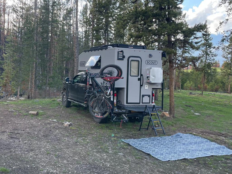  is parked on gravel at a scenic lakeside campsite.|Grey 4x4 pickup with a white pop-top Scout truck camper. A versatile RV rental for off-road camping and overland travel.|An rv rental with a grey truck camper on a pickup — photo 4