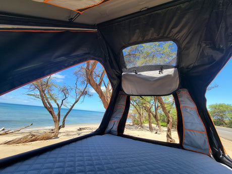 Experience beach camping with this RV rental! View from a rooftop tent: quilted mattress, blue ocean, sandy beach, and trees.