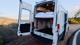 RV rental camper van with open rear doors revealing a bed & storage, parked overlooking mountains for an outdoor adventure.