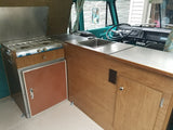 This vintage RV rental interior features a functional kitchen with a two-burner stove, sink, and custom wood cabinets.