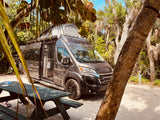 A modern gray RV rental, with its side door open and pop-top roof, is parked at a sunny, tree-lined campsite next to a picnic table.