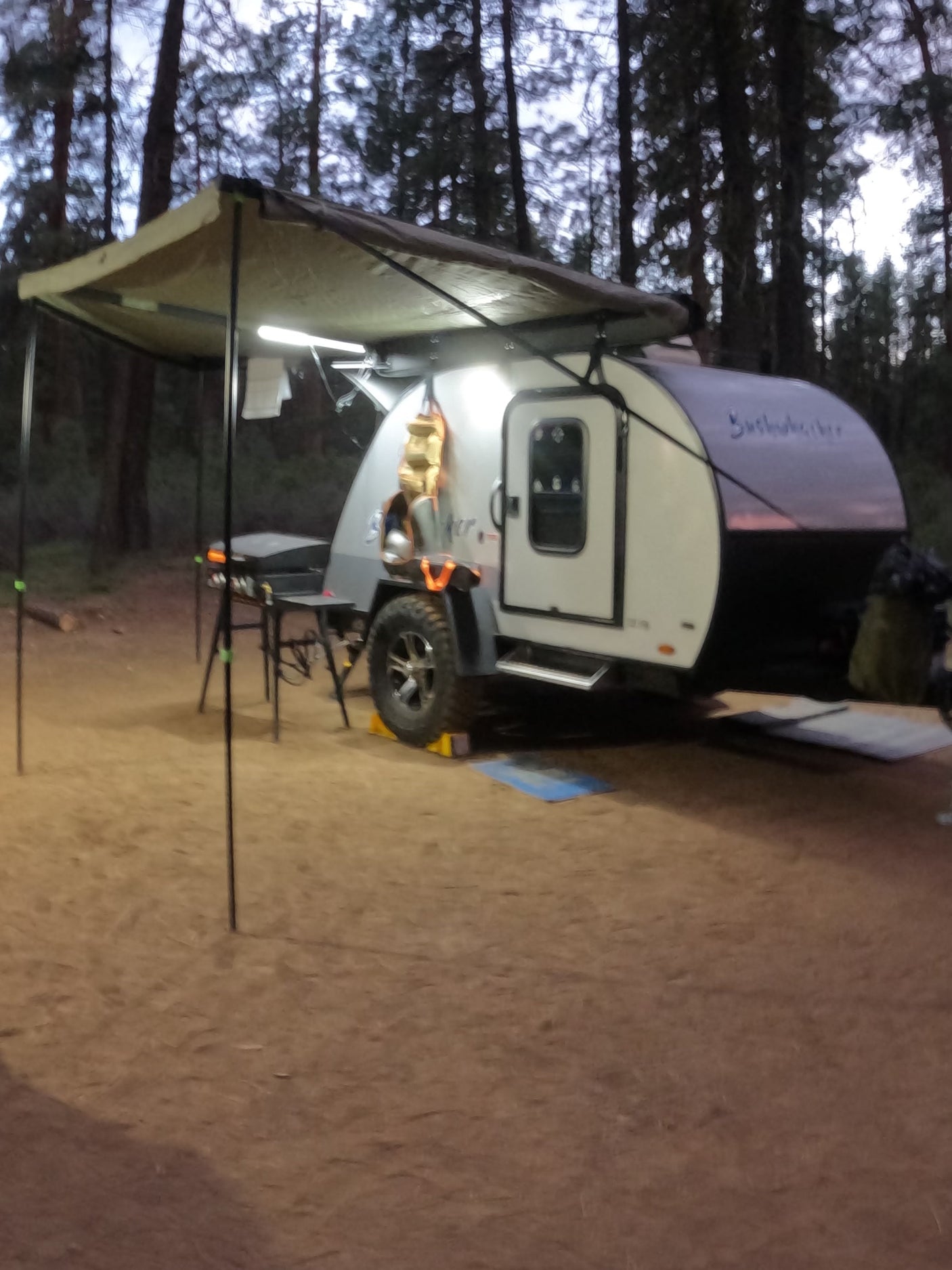 RV rental: Modern teardrop camper with illuminated awning set up for cozy forest camping adventure.