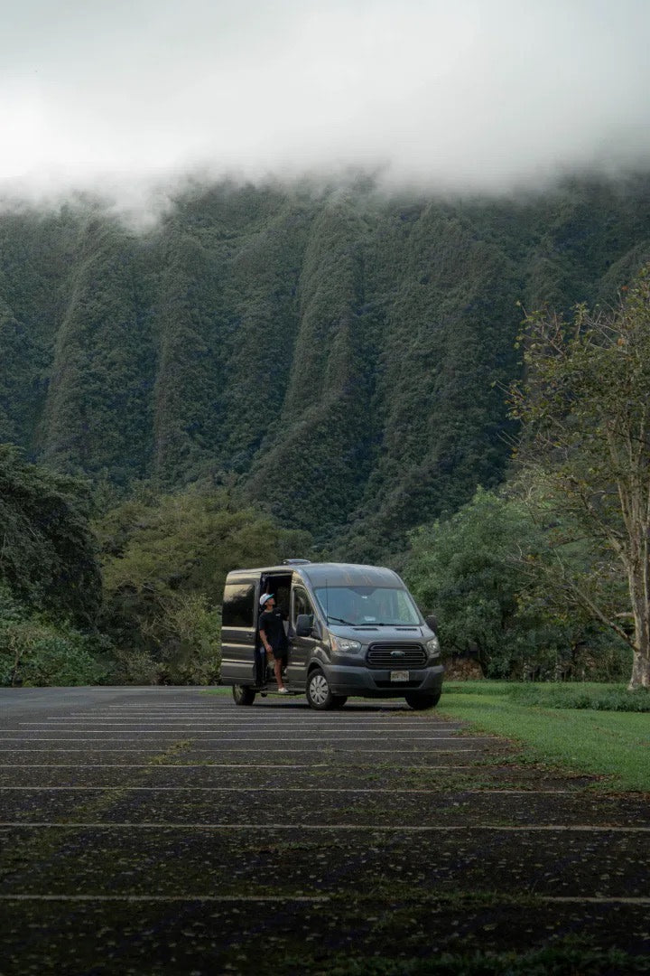 An RV rental, a grey van, is parked before lush, misty mountains. A person stands in the open door, ready for adventure.
