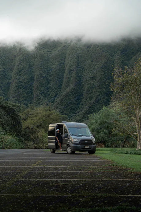 An RV rental, a grey van, is parked before lush, misty mountains. A person stands in the open door, ready for adventure.