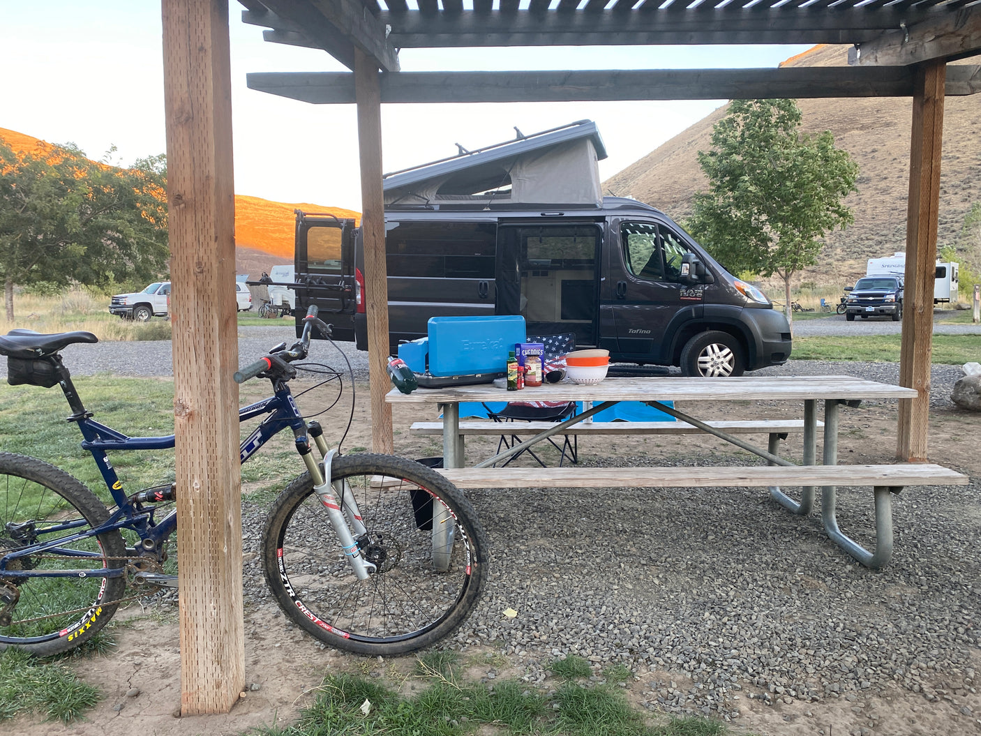 Pop-top RV rental at a serene campground with a picnic table, cooking gear, and a mountain bike. Golden hills in distance.