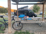 Pop-top RV rental at a serene campground with a picnic table, cooking gear, and a mountain bike. Golden hills in distance.