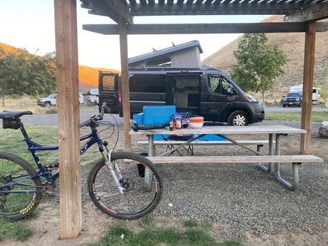 Pop-top RV rental at a serene campground with a picnic table, cooking gear, and a mountain bike. Golden hills in distance.