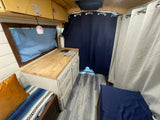 Interior of an RV rental, featuring a compact kitchenette with wood counters, a sink, and cabinets. Cozy bench seating with a colorful blanket, privacy curtains, and overhead storage create a functional living space.