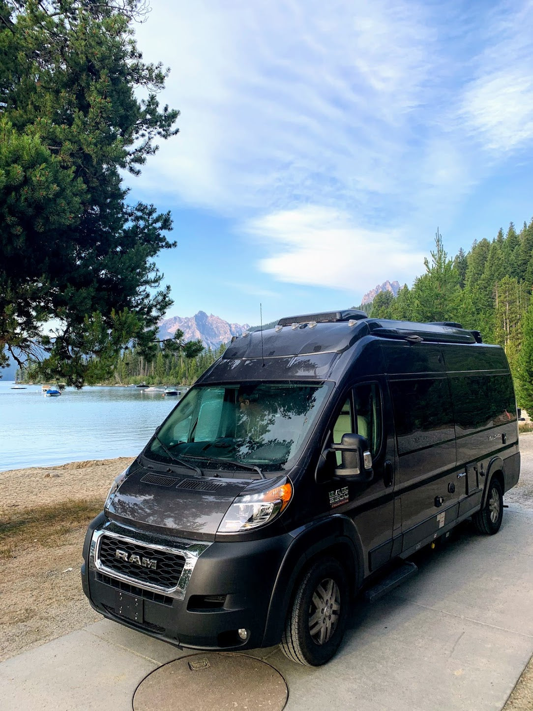 Scenic RV rental adventure! Black camper van parked by a serene lake with mountains & trees under a blue sky.
