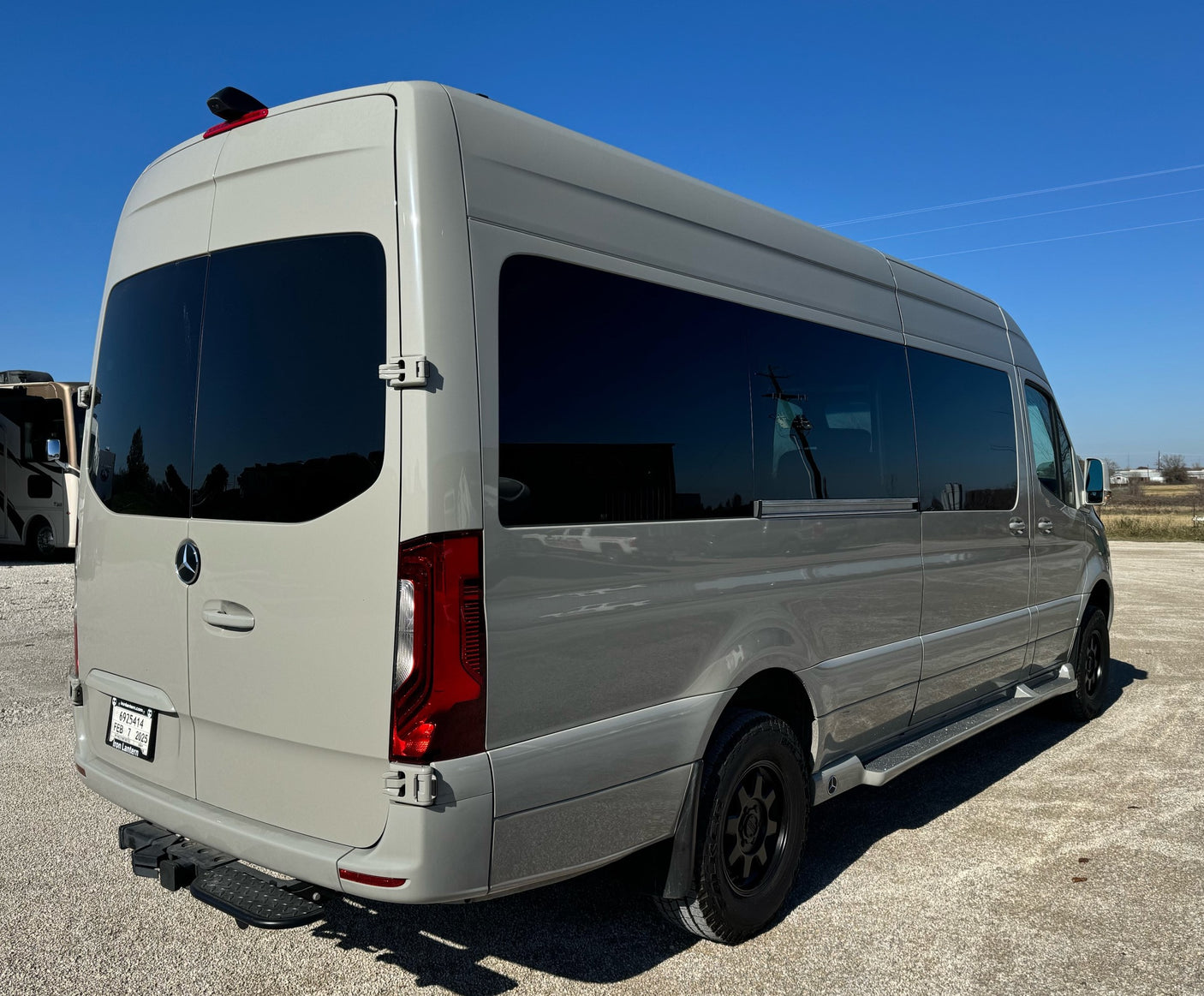 Gray Mercedes-Benz Sprinter van, perfect for rv rental trips. Features tinted windows, rear step. Parked on gravel under blue sky.