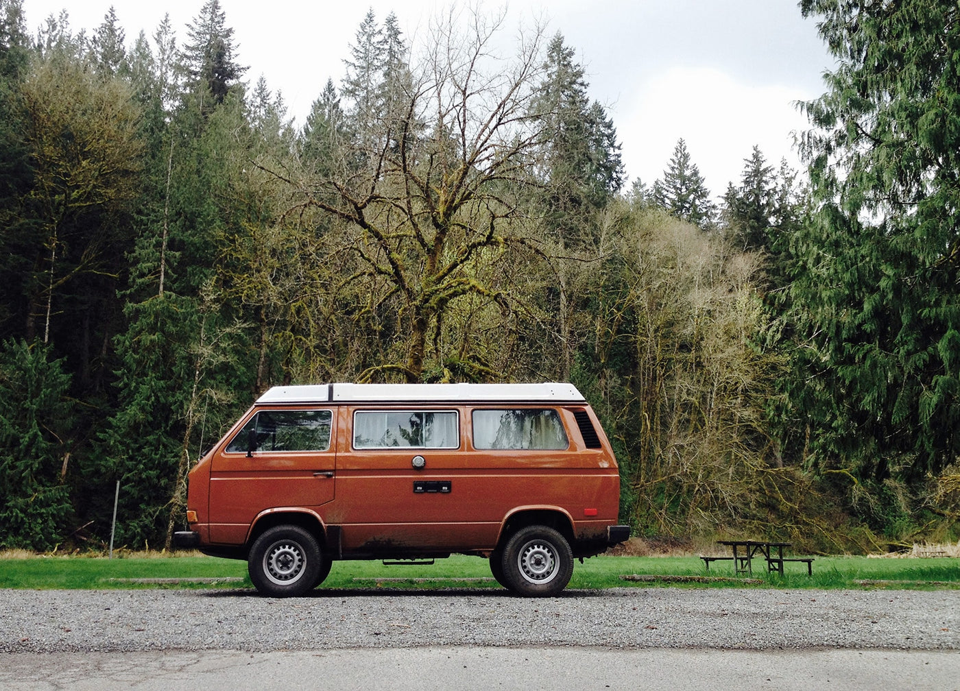 An iconic brown VW Vanagon pop-top rv rental parked on gravel. Surrounded by a dense forest & a picnic table for outdoor adventures. — photo 2