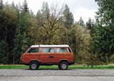 An iconic brown VW Vanagon pop-top rv rental parked on gravel. Surrounded by a dense forest & a picnic table for outdoor adventures.