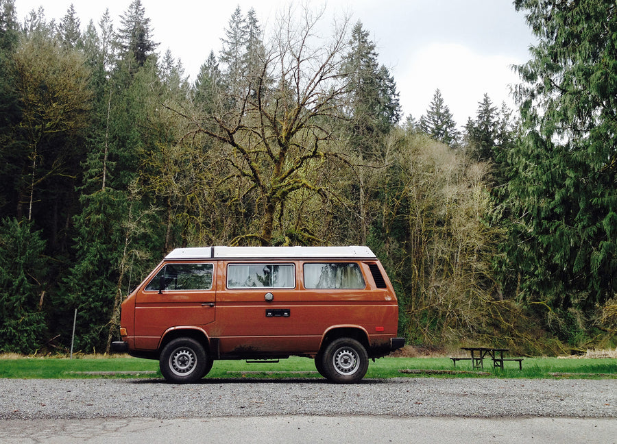 An iconic brown VW Vanagon pop-top rv rental parked on gravel. Surrounded by a dense forest & a picnic table for outdoor adventures.