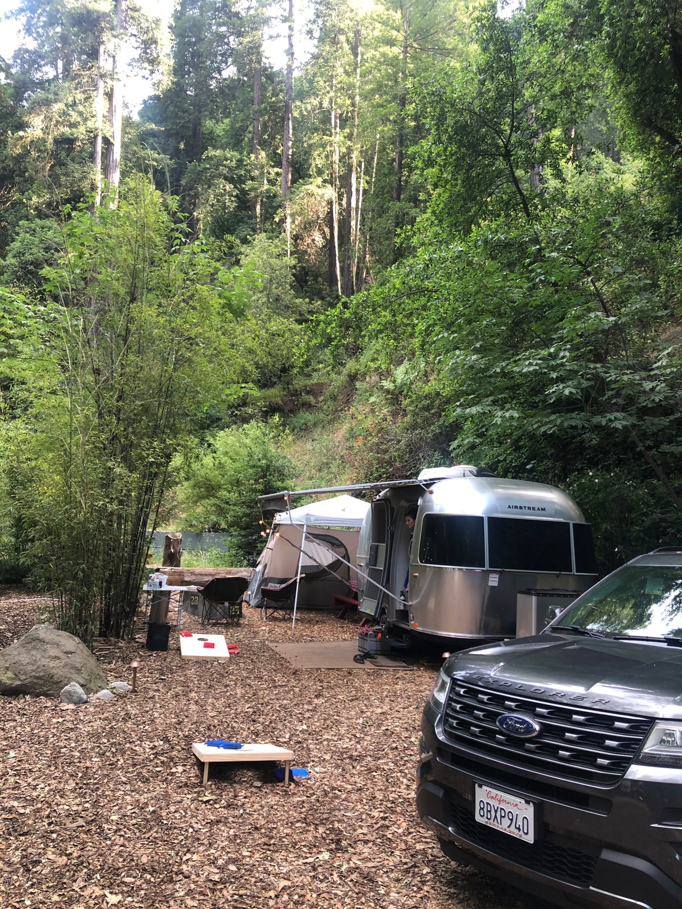 RV rental Airstream & Ford Explorer parked in a lush forest campsite, complete with a tent & cornhole boards.