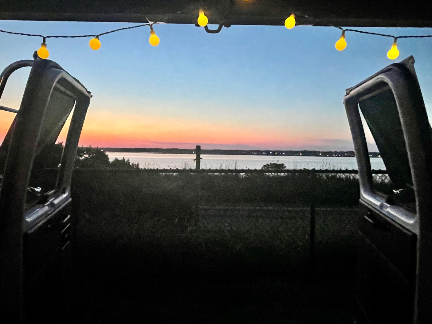 RV rental: Sunset over water, seen from open rear doors with warm string lights above.