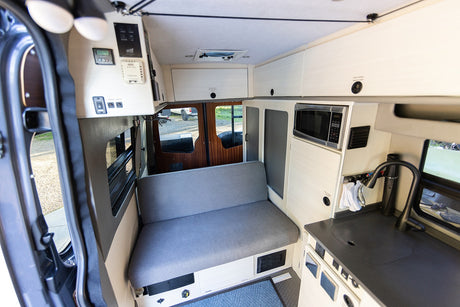 Cozy rv rental interior: A gray bench seat, compact kitchen with sink & microwave, overhead storage, & control panel.