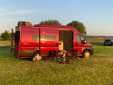This red Winnebago Solis RV rental offers a perfect camping setup. Parked in a field at sunset, with chairs, table, and a dog.
