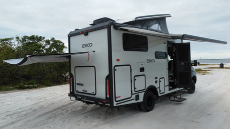 This modern Winnebago EKKO rv rental, with pop-top and extended awnings, is parked on sand near water, perfect for adventure.