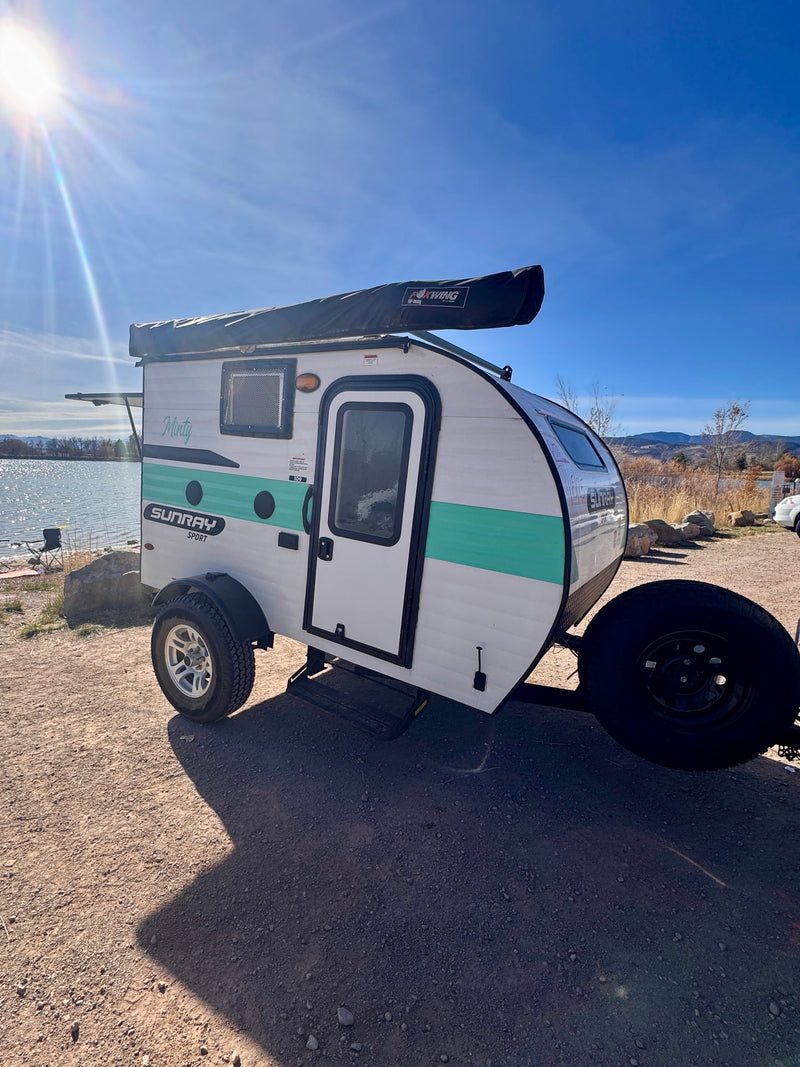 "Minty," a Sunray Sport RV rental, parked by a sunny lake with mountains. This compact travel trailer features a Foxwing awning. — photo 5