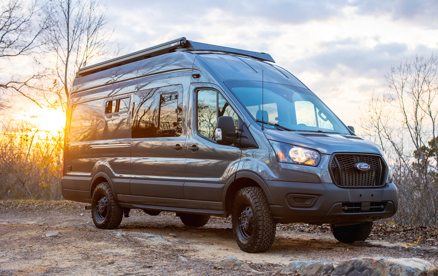 Grey Ford Transit camper van, an ideal RV rental, with off-road tires and roof awning, parked on a dirt trail at sunset.