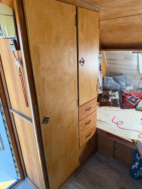 View of a vintage RV rental interior: features light wood cabinets, a cozy bed with decorative pillows, and tiled flooring.