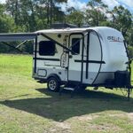 Enjoy an RV rental! A modern white and gray travel trailer, awning extended, sits on green grass with trees under a blue sky, ready for adventure.