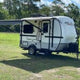 Enjoy an RV rental! A modern white and gray travel trailer, awning extended, sits on green grass with trees under a blue sky, ready for adventure.