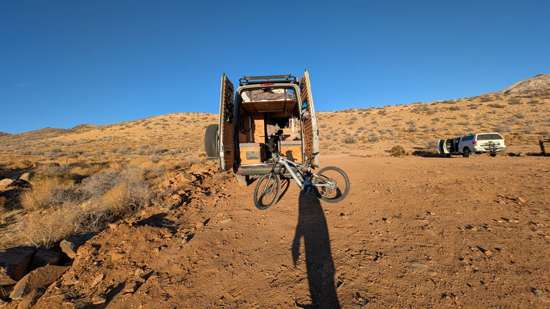 An RV rental camper van with open doors & mountain bike at camp. A person's shadow stretches across the desert ground with hills beyond. — photo 7