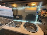 Inside an RV rental: compact kitchen with stove, sink, and glowing LED lights, framed by a serene ocean vista.