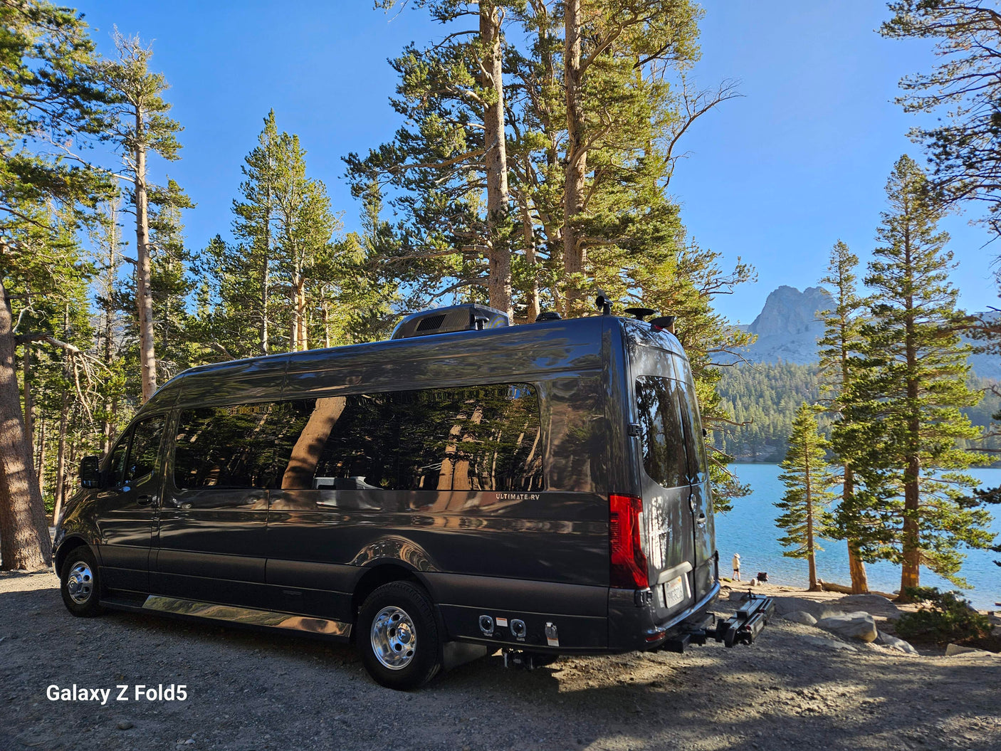 This luxurious Mercedes Sprinter RV rental is parked by a beautiful lake, surrounded by tall pine trees and mountains. Ideal for scenic adventures.