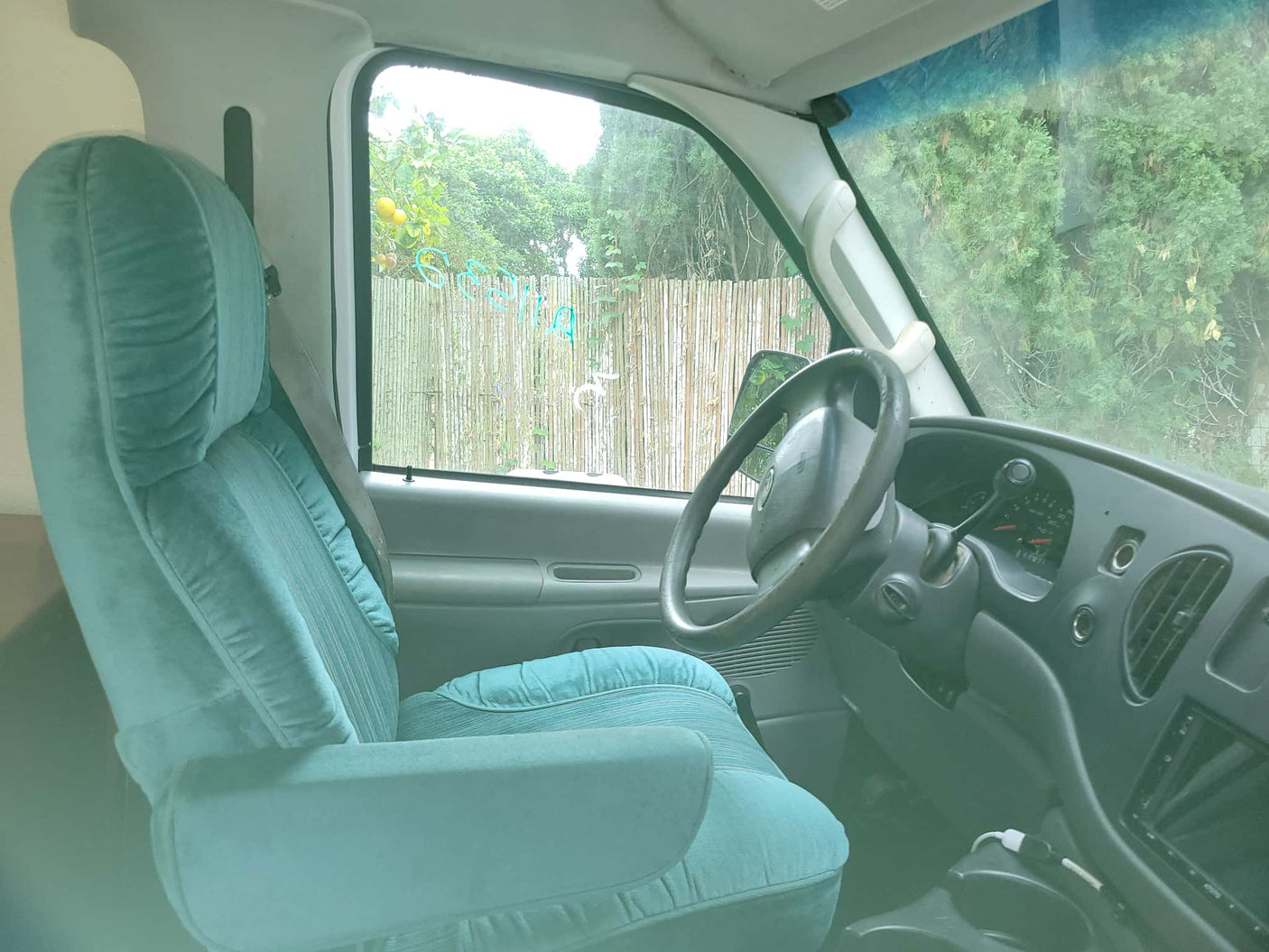 Cozy RV rental interior: teal driver's seat, dashboard, and steering wheel with lush greenery views outside.