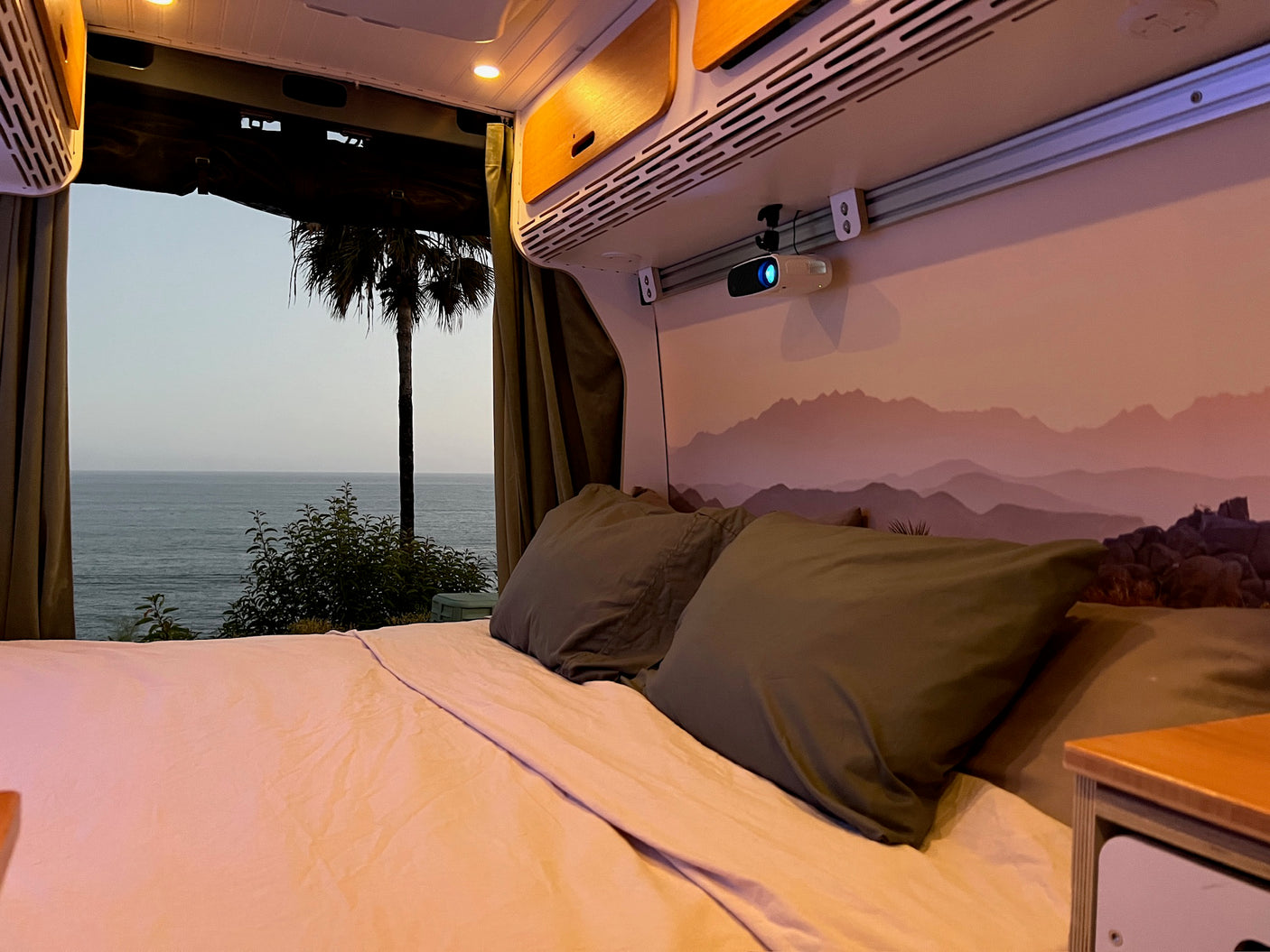Relax in this cozy RV rental bed with an ocean view and ceiling-mounted projector, offering the ultimate van life experience.