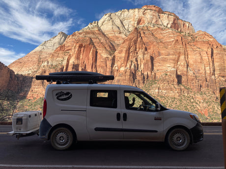 Adventure-ready RV rental van with cooler & rooftop awning, parked at iconic Zion National Park mountains. A hand waves from inside.