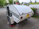 A white LittleGuy Rough Rider teardrop camper, perfect for an RV rental, with an Alaska license plate. Secured by a tire claw.