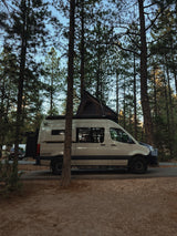 RV rental: A beige Mercedes-Benz Sprinter van with a rooftop tent, parked among tall pine trees at a serene campsite.