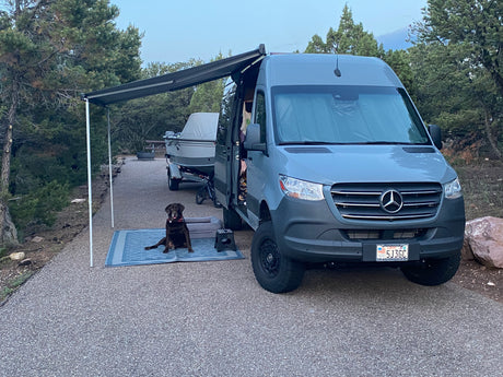 RV rental van with extended awning parked at a serene campsite. A brown dog sits on a mat, with a boat in tow.