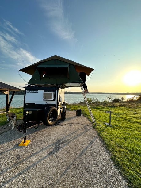 RV rental adventure: Lakeside sunset camping with a TigerMoth travel trailer, rooftop tent, and dog. Person on ladder.