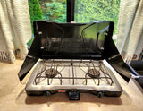 Compact kitchen setup in an RV rental: a black Coleman portable two-burner stove, lid open and ready to cook.