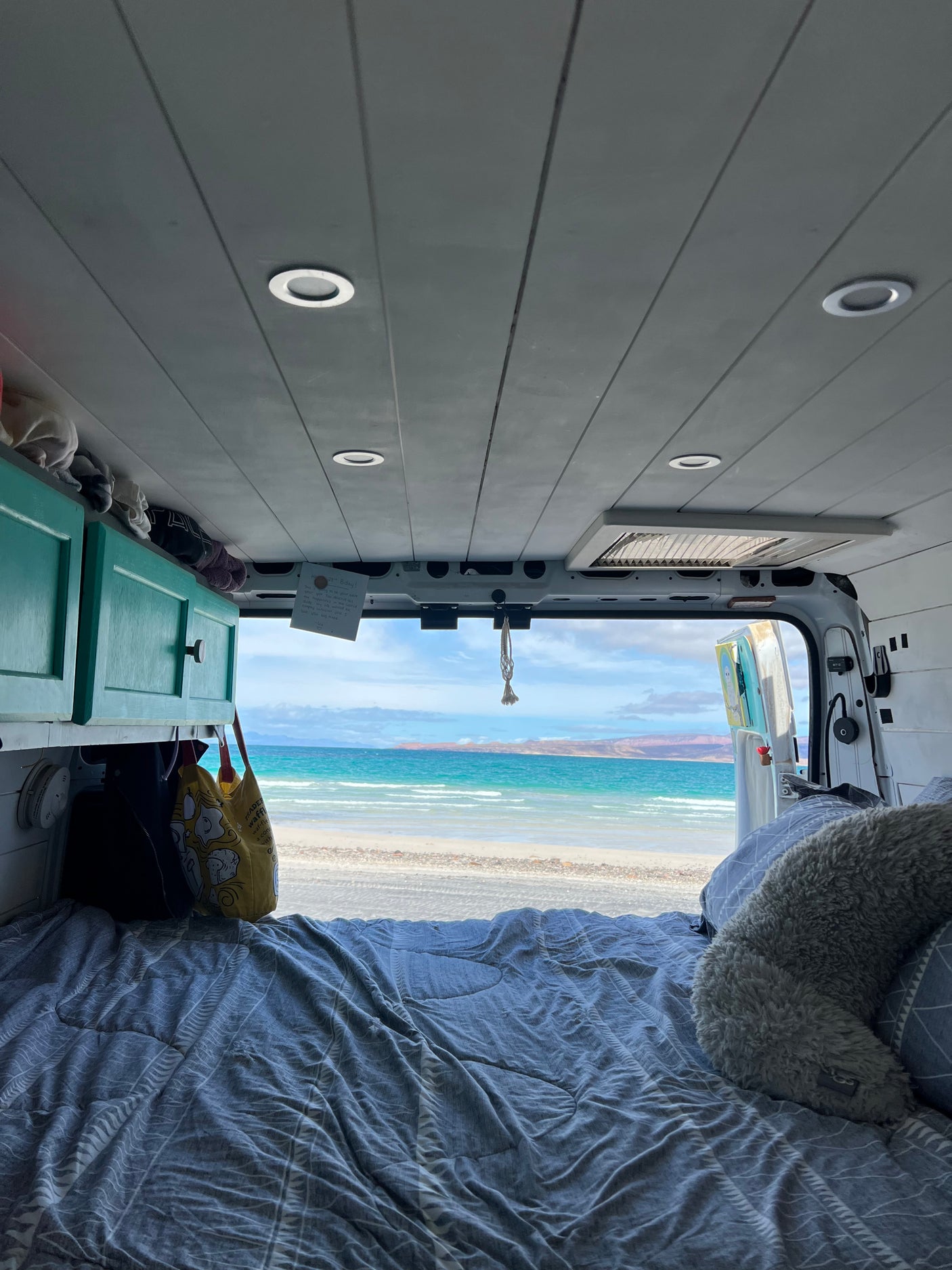 Cozy RV rental bed with a serene view of a sandy beach, turquoise ocean, and distant mountains. Enjoy the ultimate van life escape!