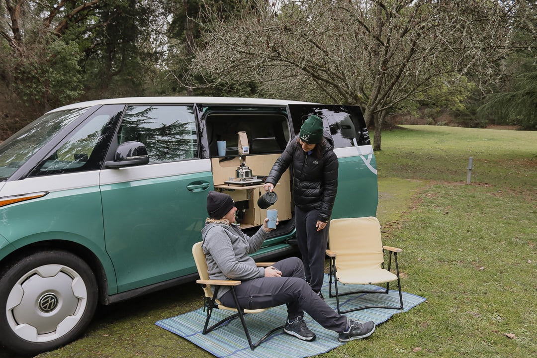 Two friends enjoy coffee outside a green/white camper van, featuring a roadside kitchen setup. Perfect for your next rv rental camping trip!