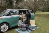 Two friends enjoy coffee outside a green/white camper van, featuring a roadside kitchen setup. Perfect for your next rv rental camping trip!