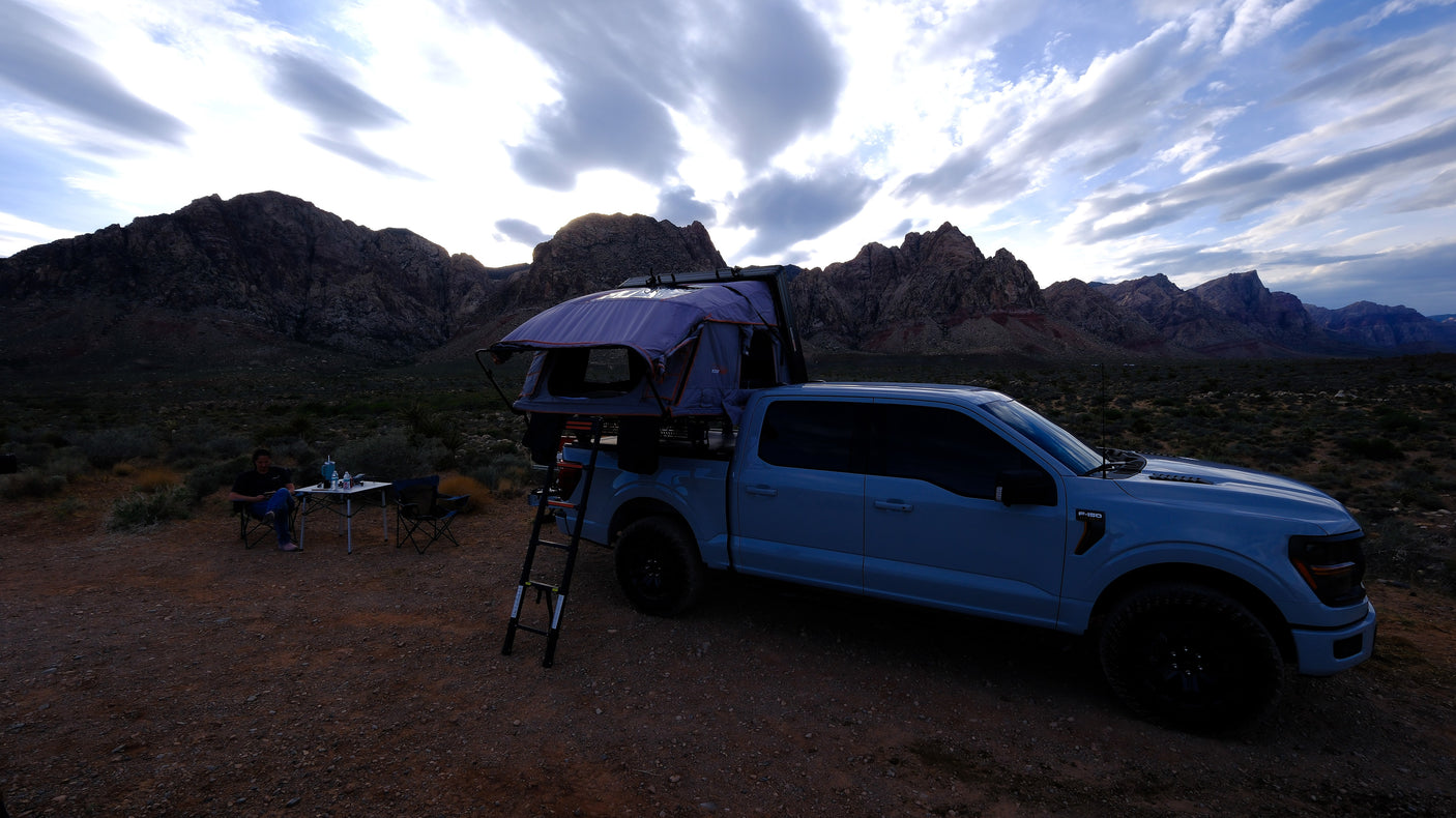Truck with rooftop tent and person camping. Mountains under cloudy skies create an epic scene. A versatile alternative to an RV rental.