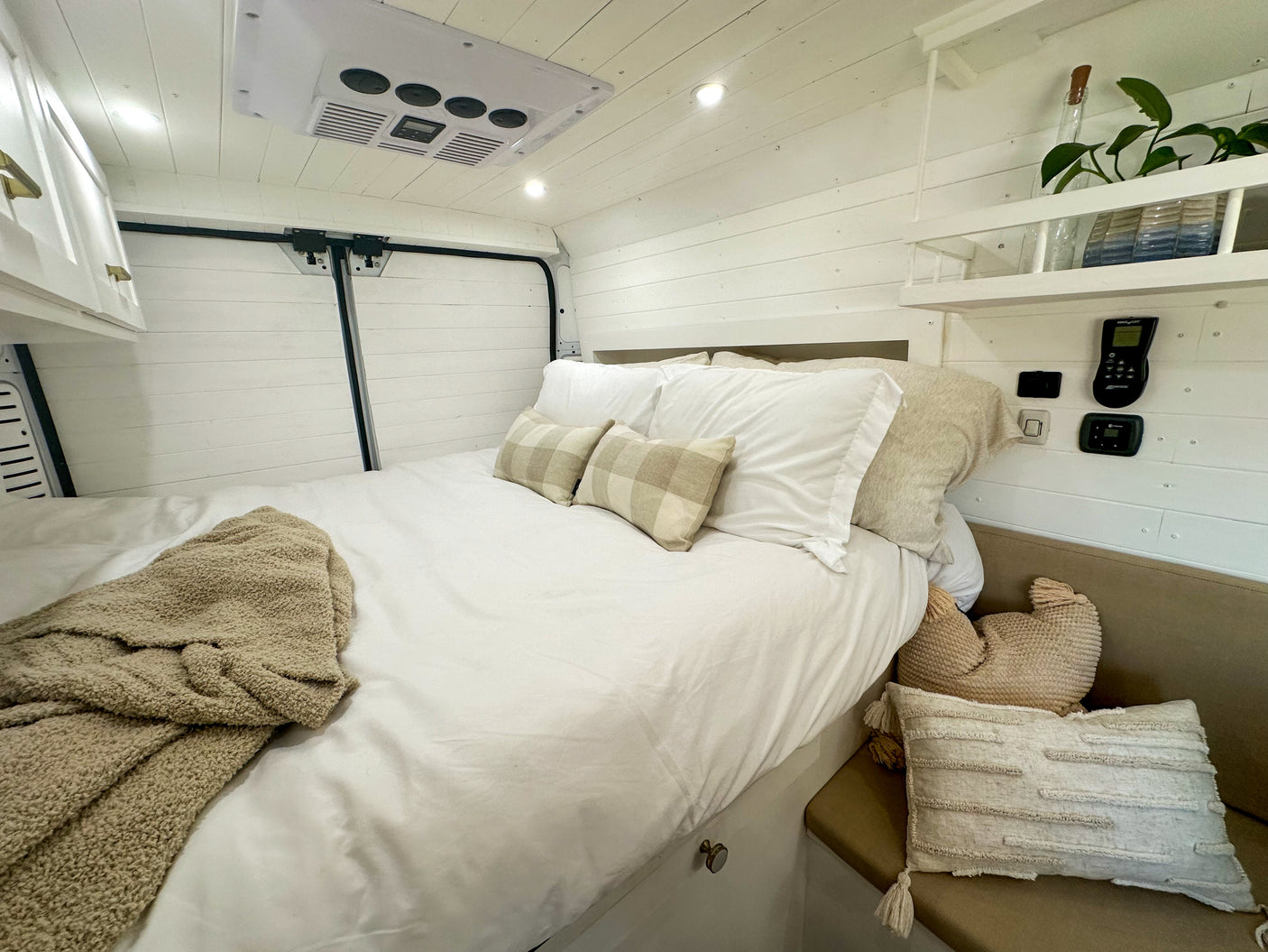 RV rental interior: A bright bedroom with white shiplap walls, soft bedding, built-in shelf, and controls.