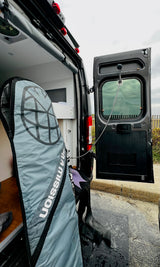 RV rental with rear doors open, revealing a surf gear bag & mobile shower. Wetsuit boots are on the wet ground.