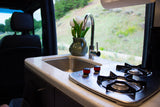 RV rental: A compact camper kitchen with a sink, faucet, two-burner stove, and white tulips by the window.