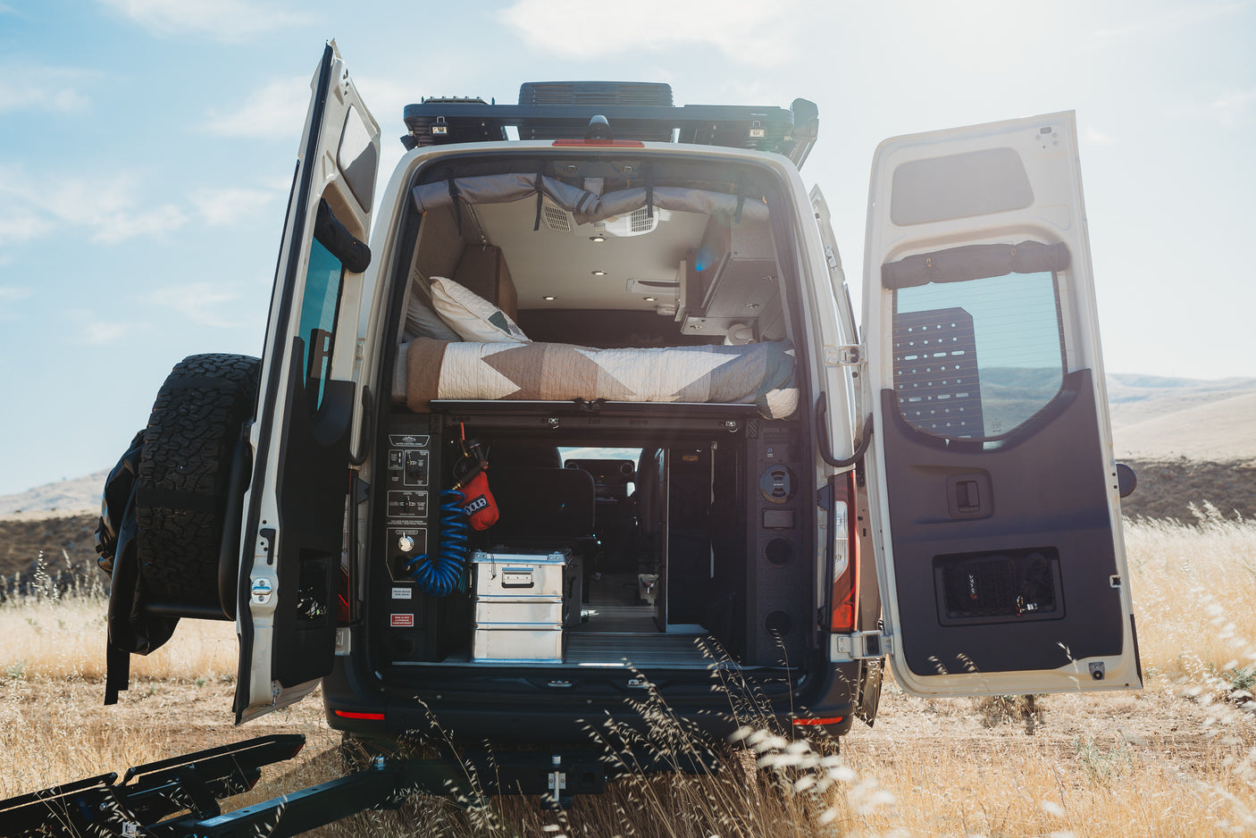 Discover this adventure-ready rv rental camper van, with its rear doors open, showing a bed, gear, and utility controls for travel.