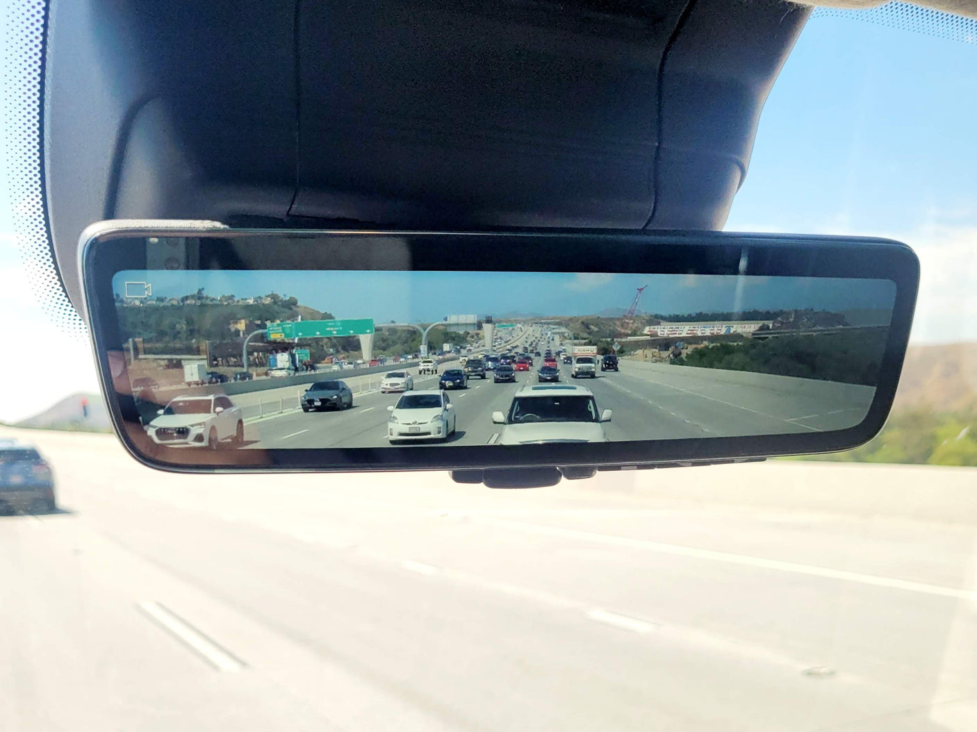 Digital rearview mirror shows live highway traffic under clear skies. A large white vehicle, possibly an RV rental, travels among cars.