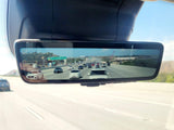 Digital rearview mirror shows live highway traffic under clear skies. A large white vehicle, possibly an RV rental, travels among cars.