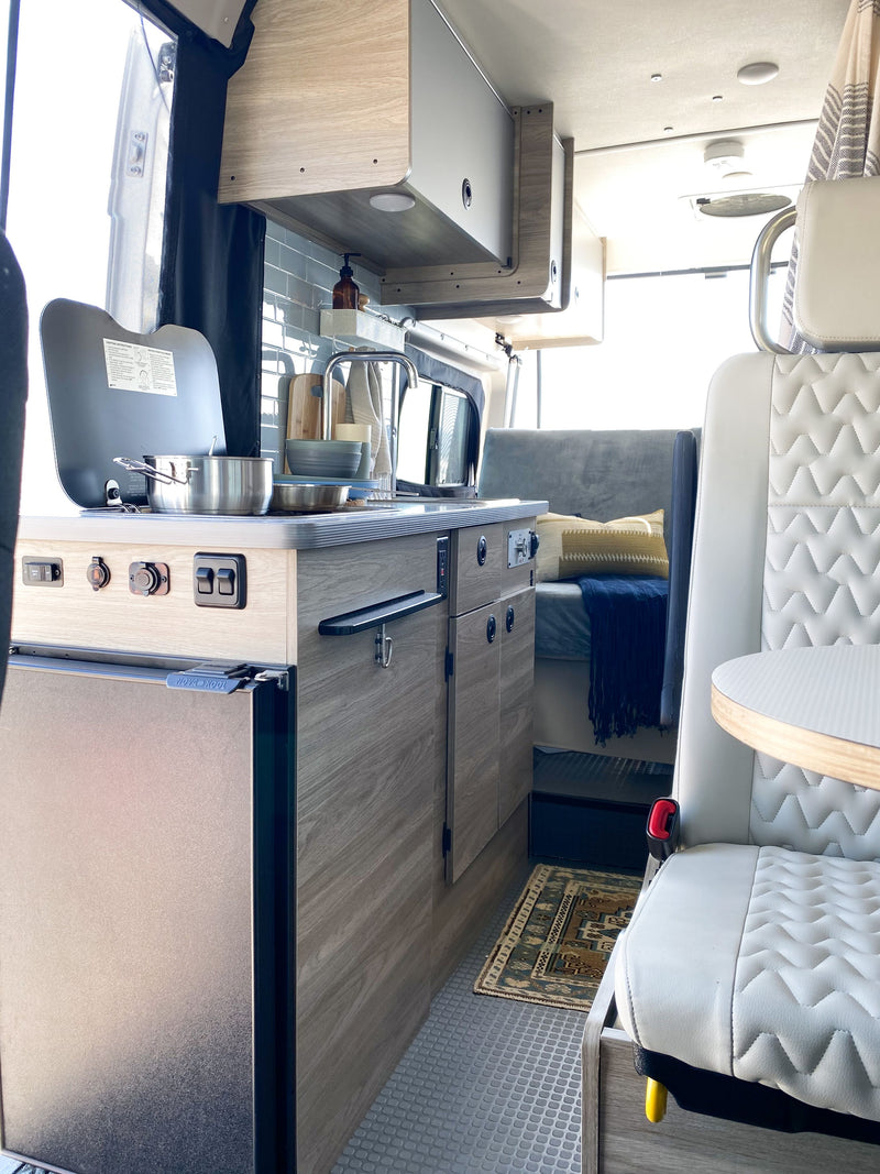 This RV rental interior features a compact kitchen with cooktop & refrigerator, seating with a table, and rear bed area. — photo 5