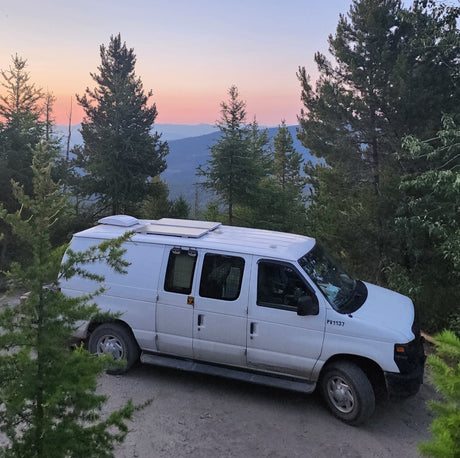 White RV rental van with solar panels on roof, parked on dirt among pines, overlooking a vibrant mountain sunset.